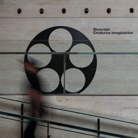 Interior shot of a person walking by an art installation featuring a film reel design and text, against a textured beige wall.