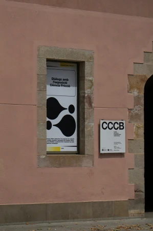 Exterior view of a pink building with posters in a window and on the adjacent wall, featuring abstract designs and text.