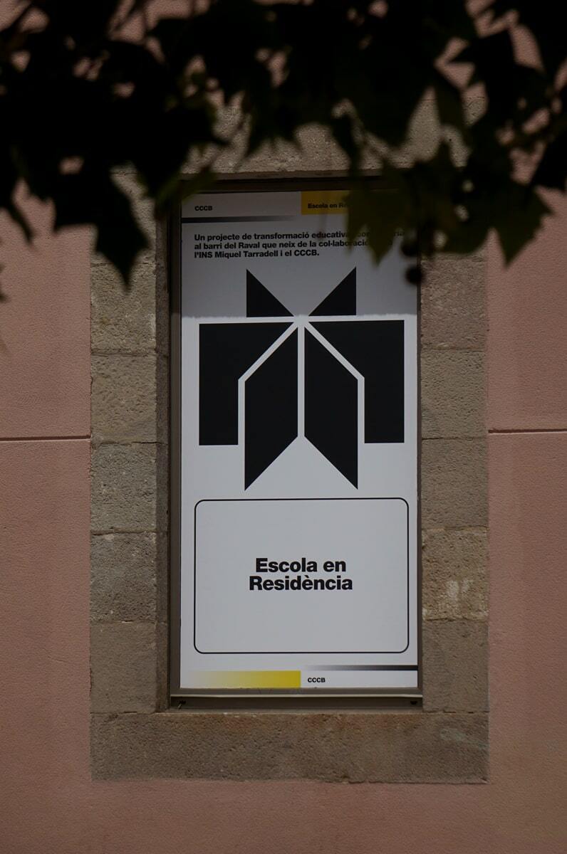 Architectural sign on a light pink stone building features a black abstract geometric figure and text in Catalan. Leaves in foreground.
