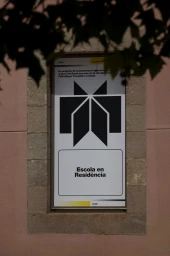 A poster featuring a black geometric logo and the text Escola en Resència mounted on a pink wall with stone framing.