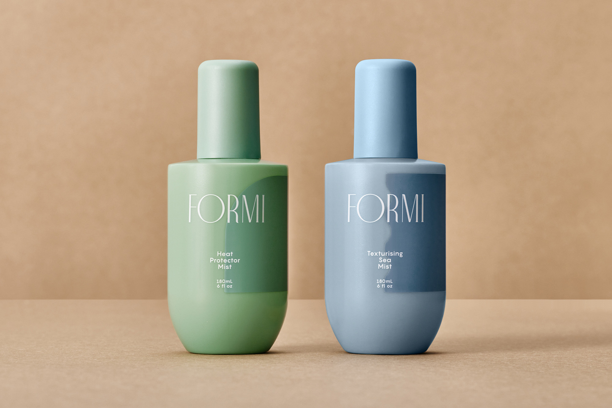 Two pastel-colored 'Formi' brand mist bottles (green heat protector, blue texturizing) sit side-by-side against a neutral studio backdrop.