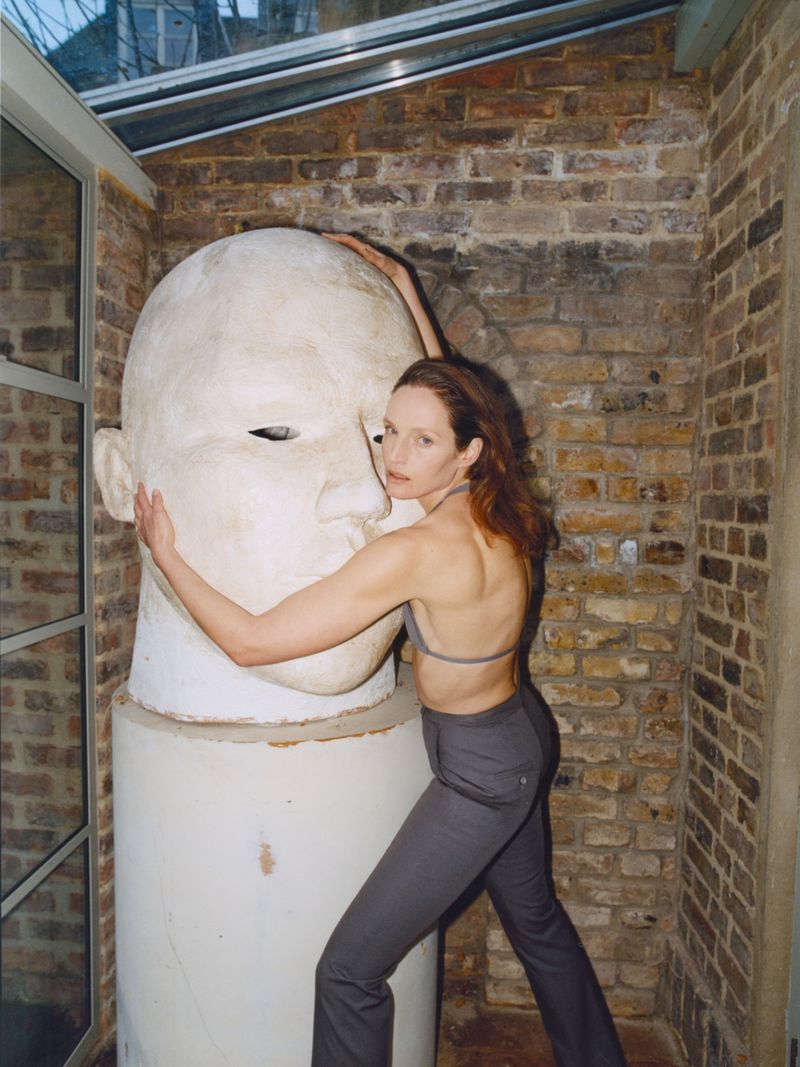 Woman in bra top and pants poses next to a large sculpture of a human head in an indoor setting with brick walls and natural light.