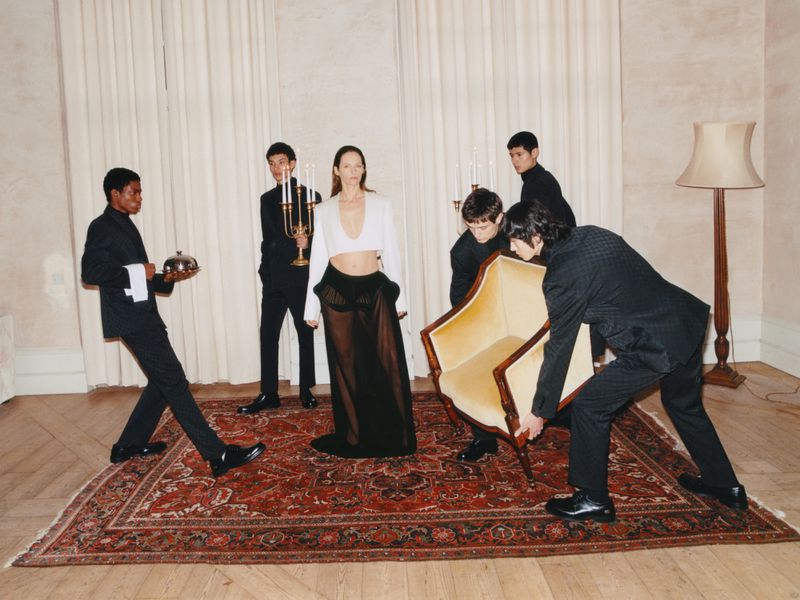 Editorial fashion shot of a model surrounded by a group of people in a room with a patterned rug and ornate wall decor, indoor setting.