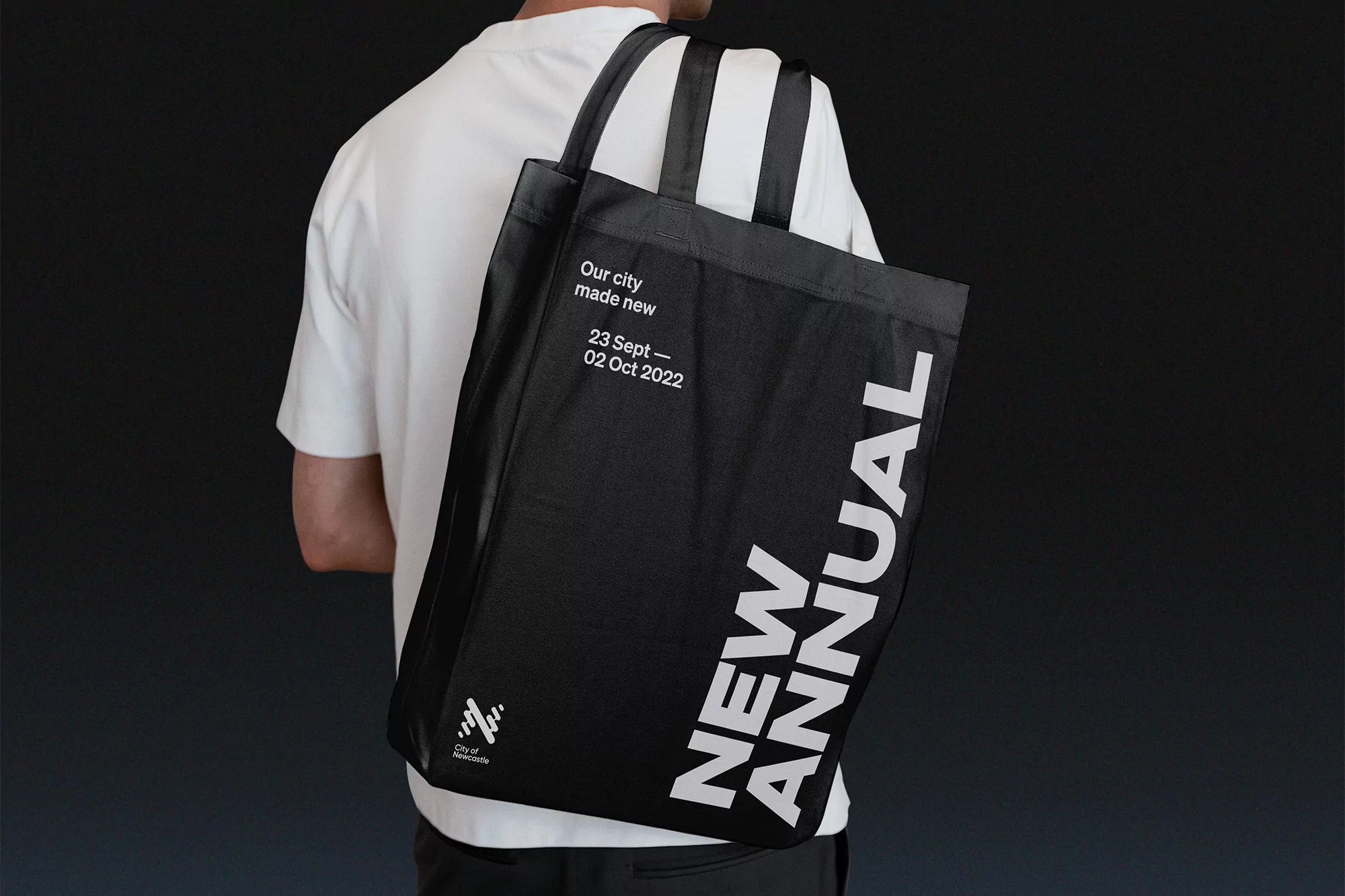 Person wearing a white t-shirt carrying a large black tote bag with bold white New Annual typography on a dark background.