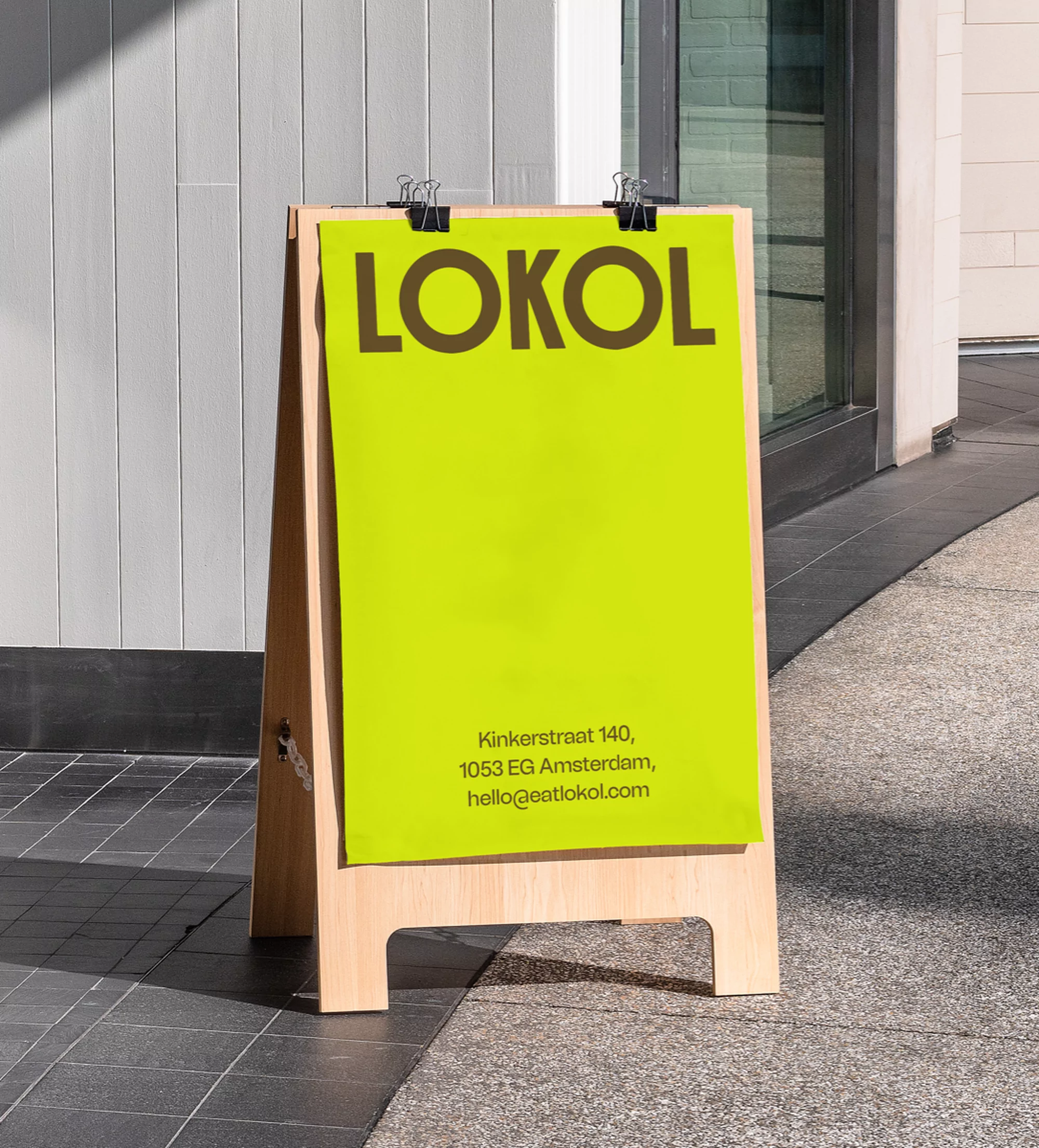 Sidewalk sign with wooden frame and bright yellow poster. Sign reads 'LOKOL' with address information displayed on the lower portion.
