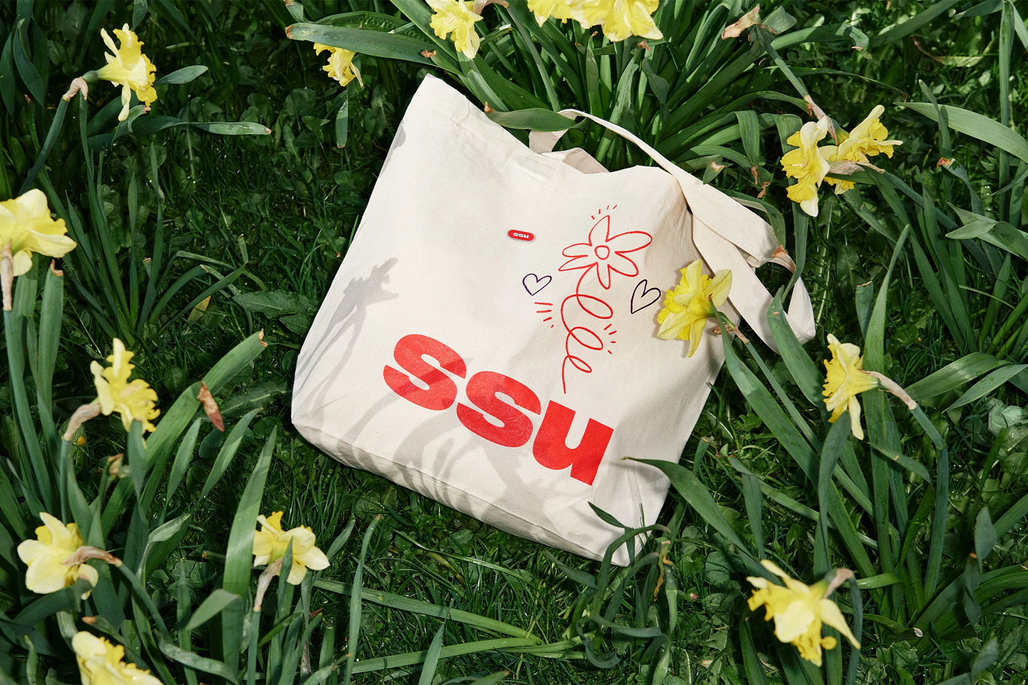 An ecru tote bag with red graphic design sits on a bed of green grass dotted with yellow daffodils on a sunny day.
