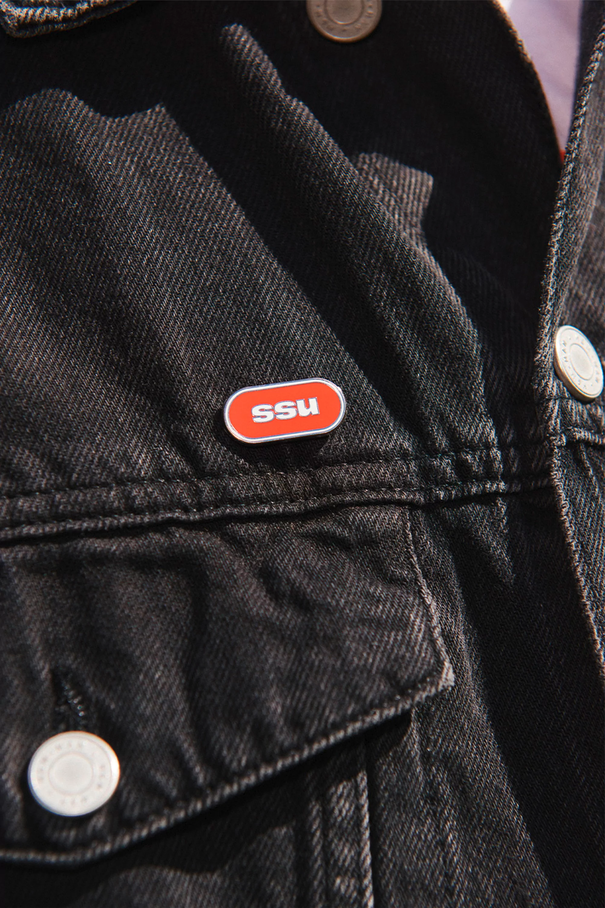 Close-up of a black denim jacket with an orange pin on the lapel, focus on the texture and color contrast of the jacket and pin.