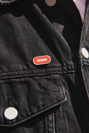 Close-up of a black denim jacket with an orange pin on the lapel, focus on the texture and color contrast of the jacket and pin.