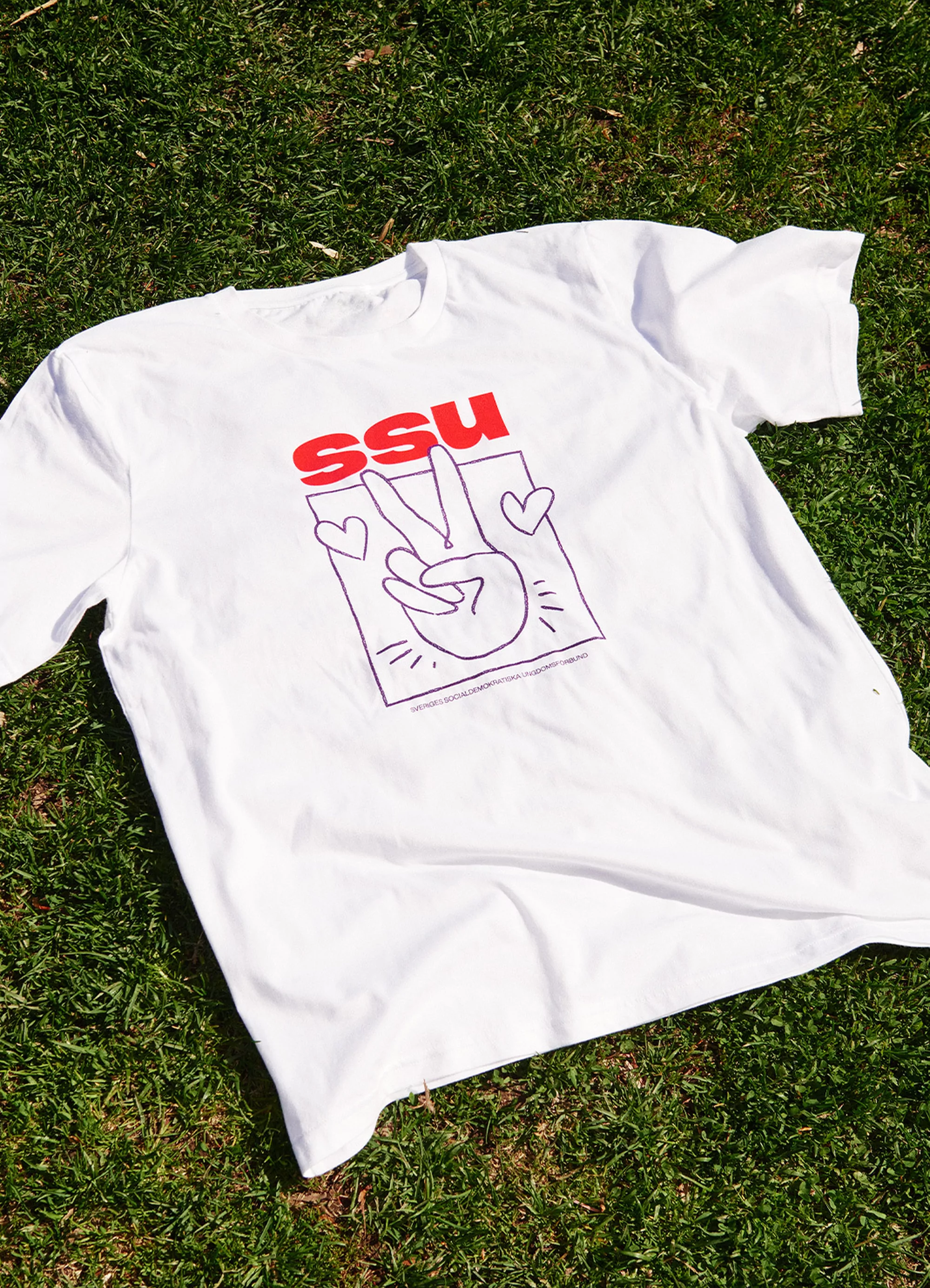 White t-shirt with a hand giving the peace sign design and the letters SSU lying on green grass in an outdoor setting.