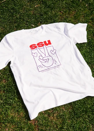 White t-shirt with a hand giving the peace sign design and the letters SSU lying on green grass in an outdoor setting.