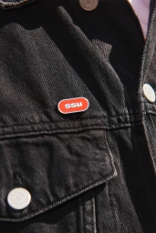 Close-up of a black denim jacket pocket featuring a small red and white ssu enamel pin for the brand identity project by Martina Huber.