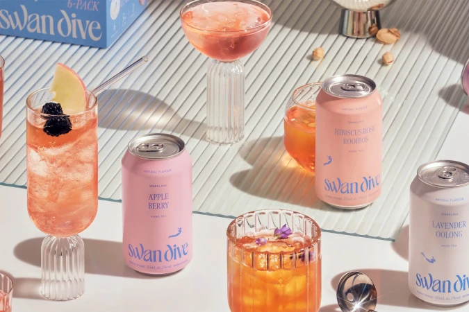 Several colorful cans and glasses of Swan Dive hard tea beverages on a table with a light blue striped background, close-up product shot.