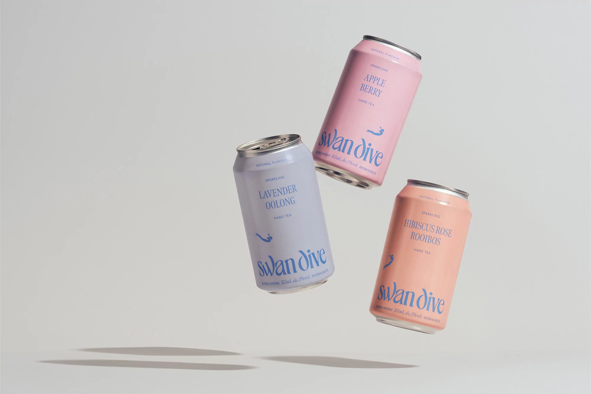 Three colorful cans of Swan Dive sparkling tea float in a simple studio setting. The pastel-colored cans are pink, lavender and coral.