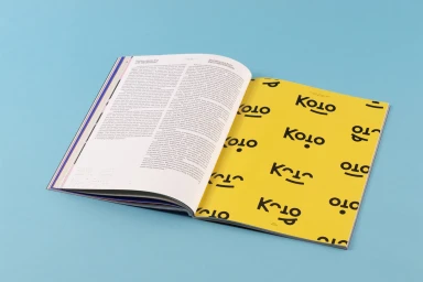 An open magazine lies on a light blue surface, showing printed text on one page and bold typographic patterns on a bright yellow page.