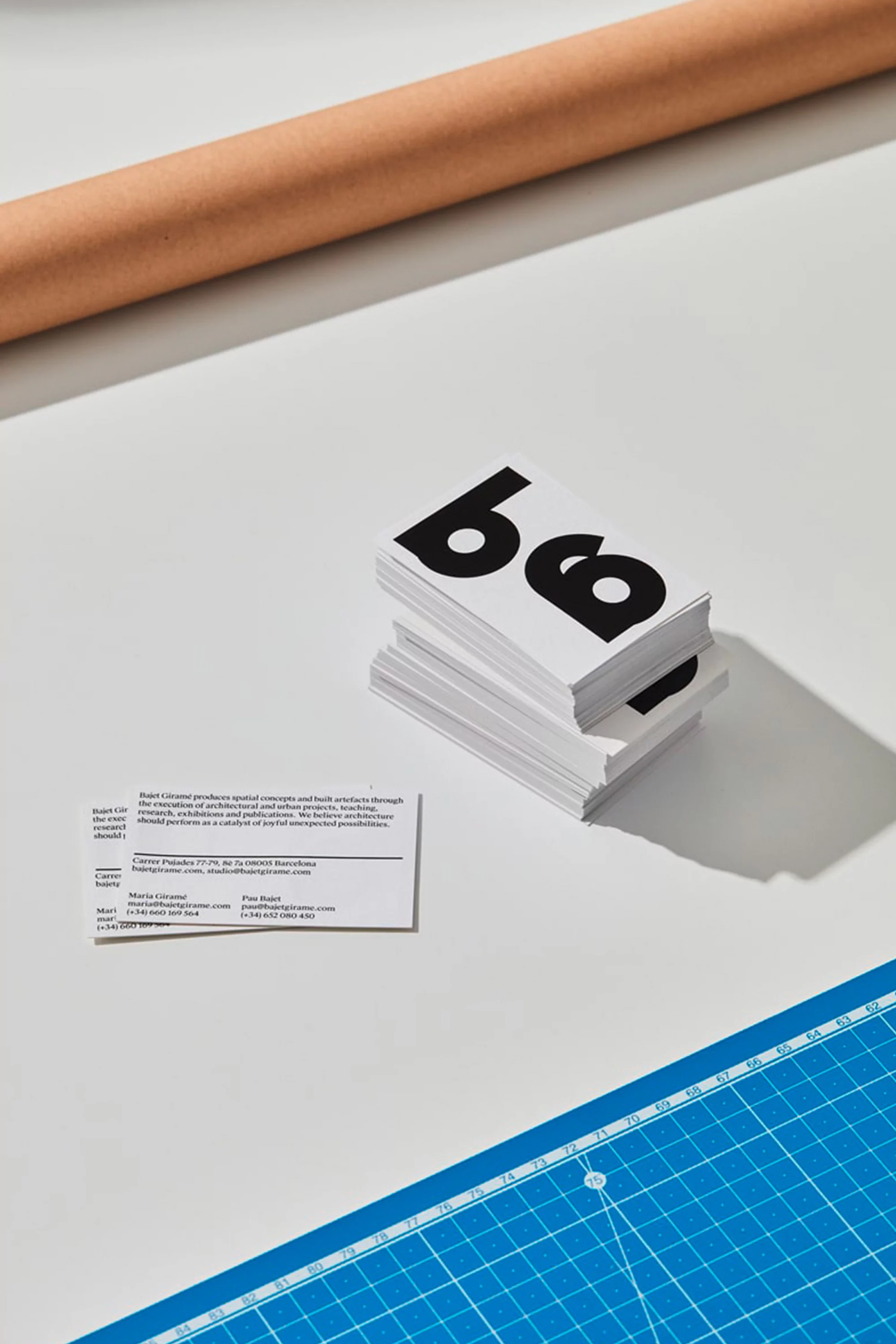 Stack of business cards with bold typography and contact information, placed on a white surface with a brown tube and blue cutting mat.