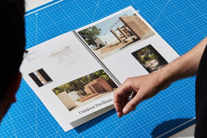 Open design book on a blue grid cutting mat, featuring architectural renderings and photographs of outdoor pavilions, showcasing outdoor architecture.
