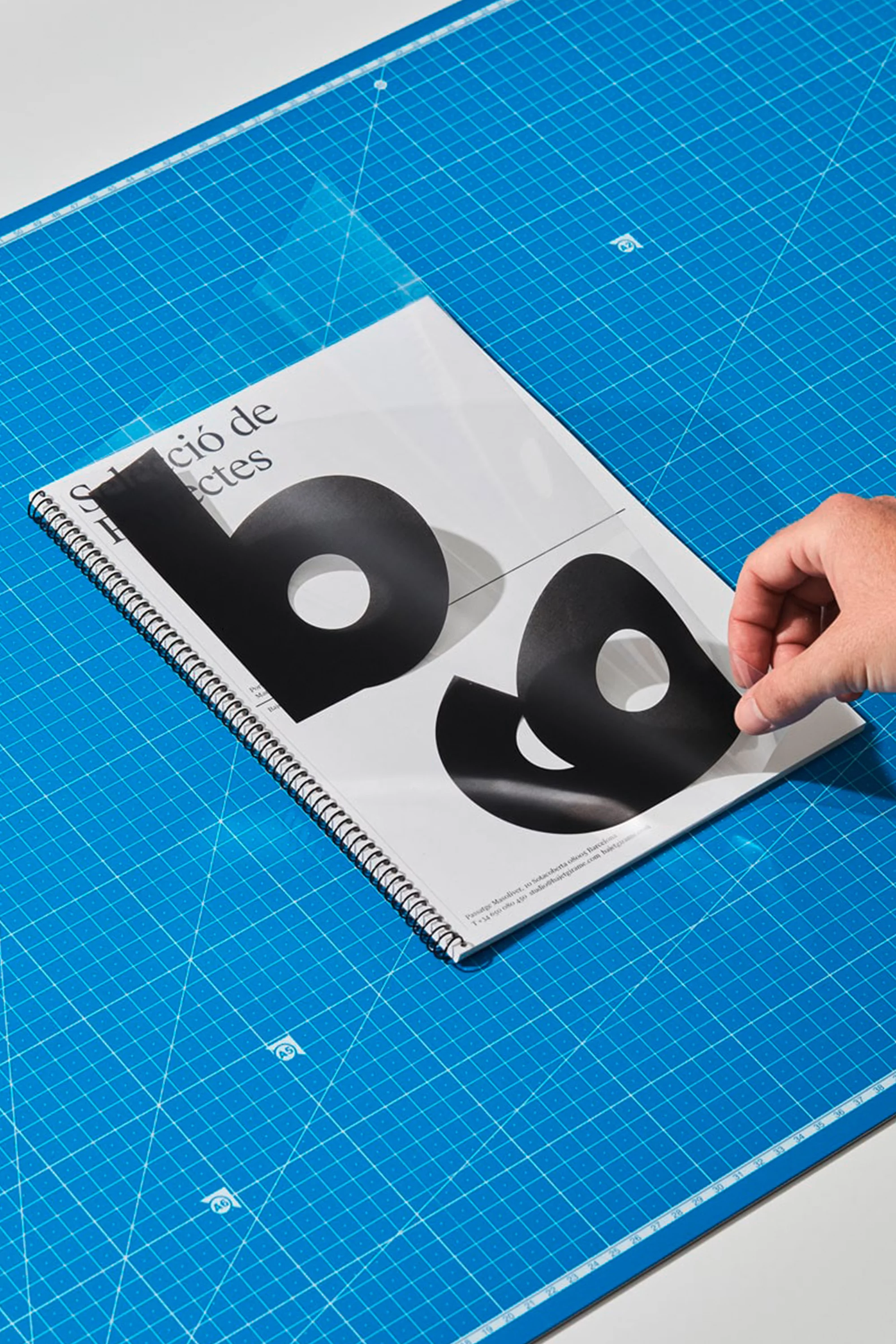 Close-up of a person's hand placing large black graphic letters on a white spiral-bound notebook over a blue grid cutting mat.