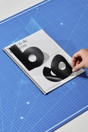 Hand peeling a transparent overlay off a spiral-bound notebook with bold typography resting on a blue cutting mat
