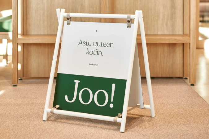 A-frame sign stands on a neutral carpet, displaying promotional text in Finnish on a white and green background with light wood backdrop.