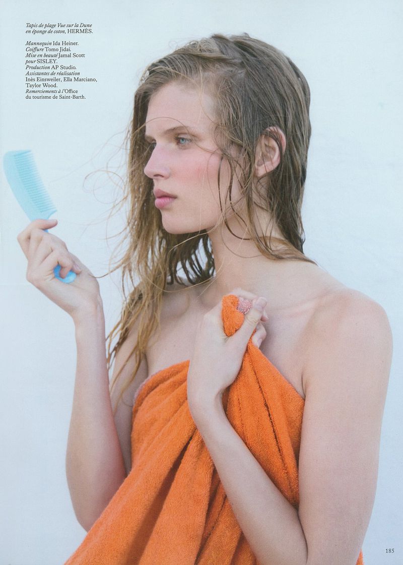 Portrait of a fair-skinned woman with wet hair, wrapped in an orange towel, holding a blue comb, against a light-colored background.