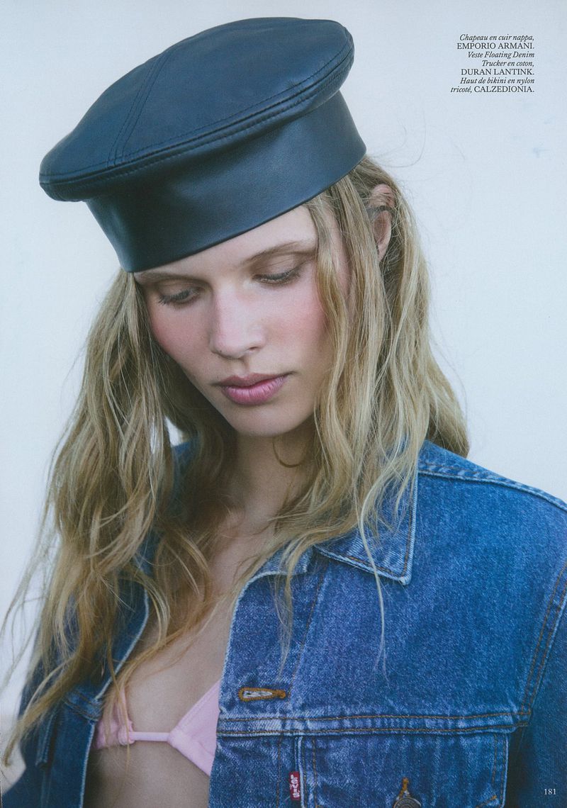 Portrait of a young woman with wavy blonde hair wearing a dark blue leather beret and a denim jacket against a white background.