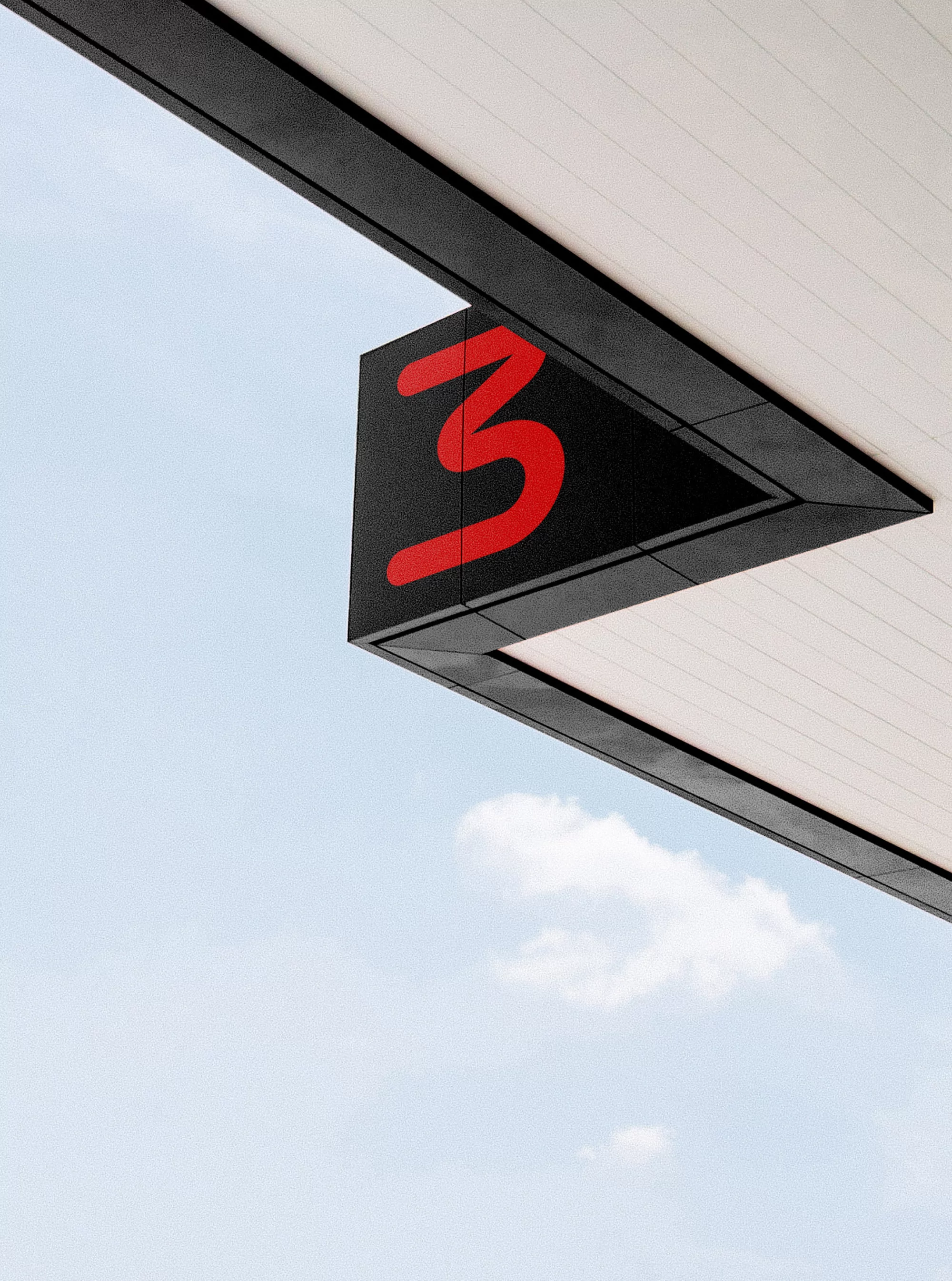 A black triangular sign displaying a red number three mounted underneath a white architectural overhang against a clear blue sky