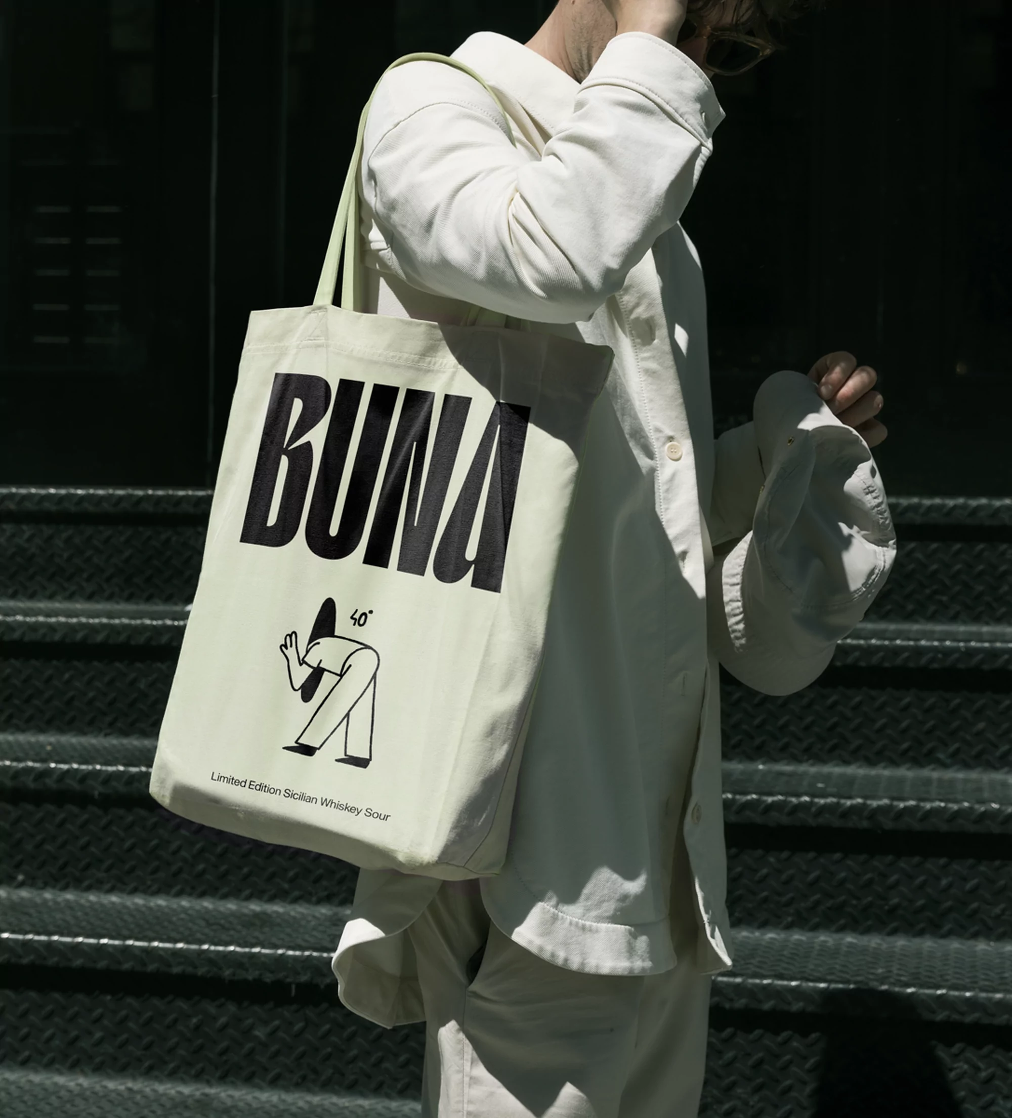 Person in white outfit carries a canvas tote bag with a cartoon graphic and the word 'BUNA' printed on it while standing outdoors.