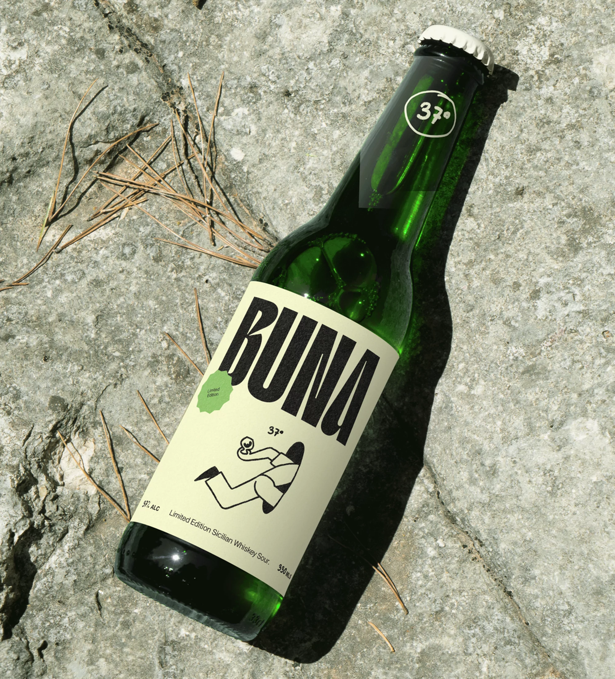 A green glass bottle of Buina Limited Edition Sicilian Whiskey Sour rests on a textured rock surface with scattered pine needles.