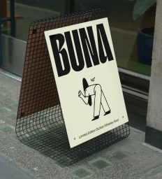 A-frame sidewalk sign featuring the BUNA brand identity with bold black typography and minimalist character illustration.