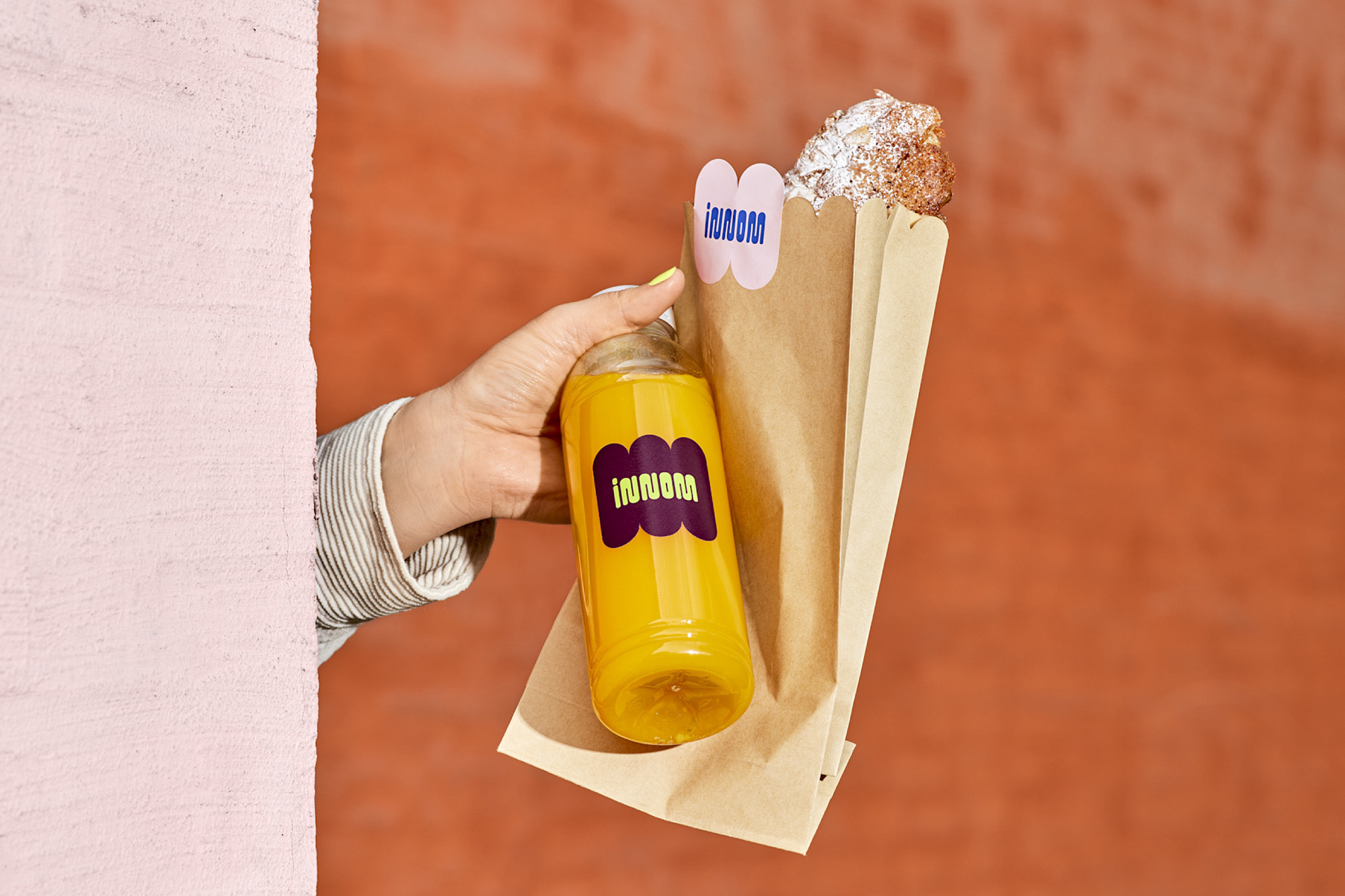 Hand holding an orange juice bottle and a paper bag with food against a coral wall, featuring a minimalist logo design.