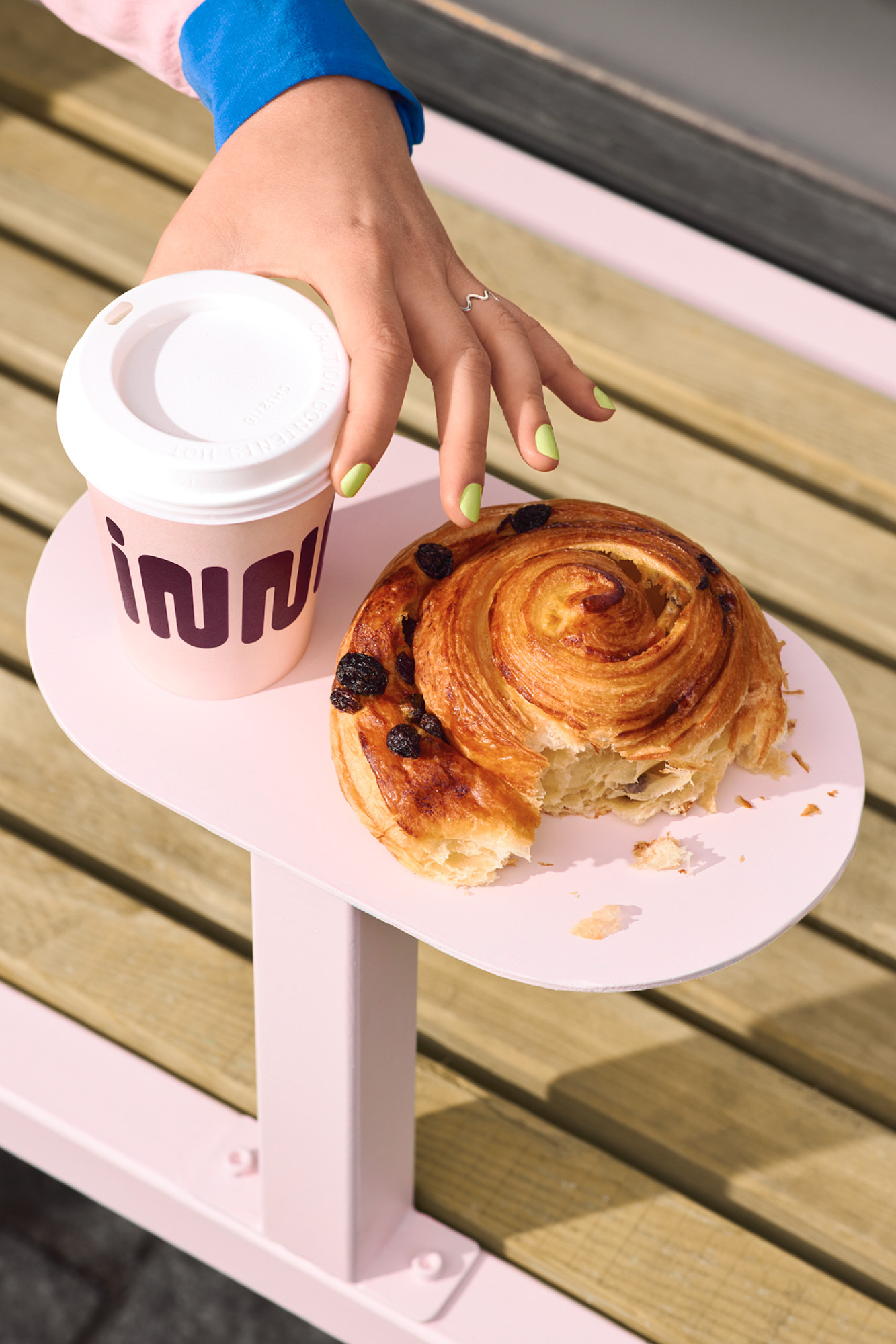 A hand reaches for a pastry and a coffee cup on a small pink table outside, with slatted wood seating in the background.