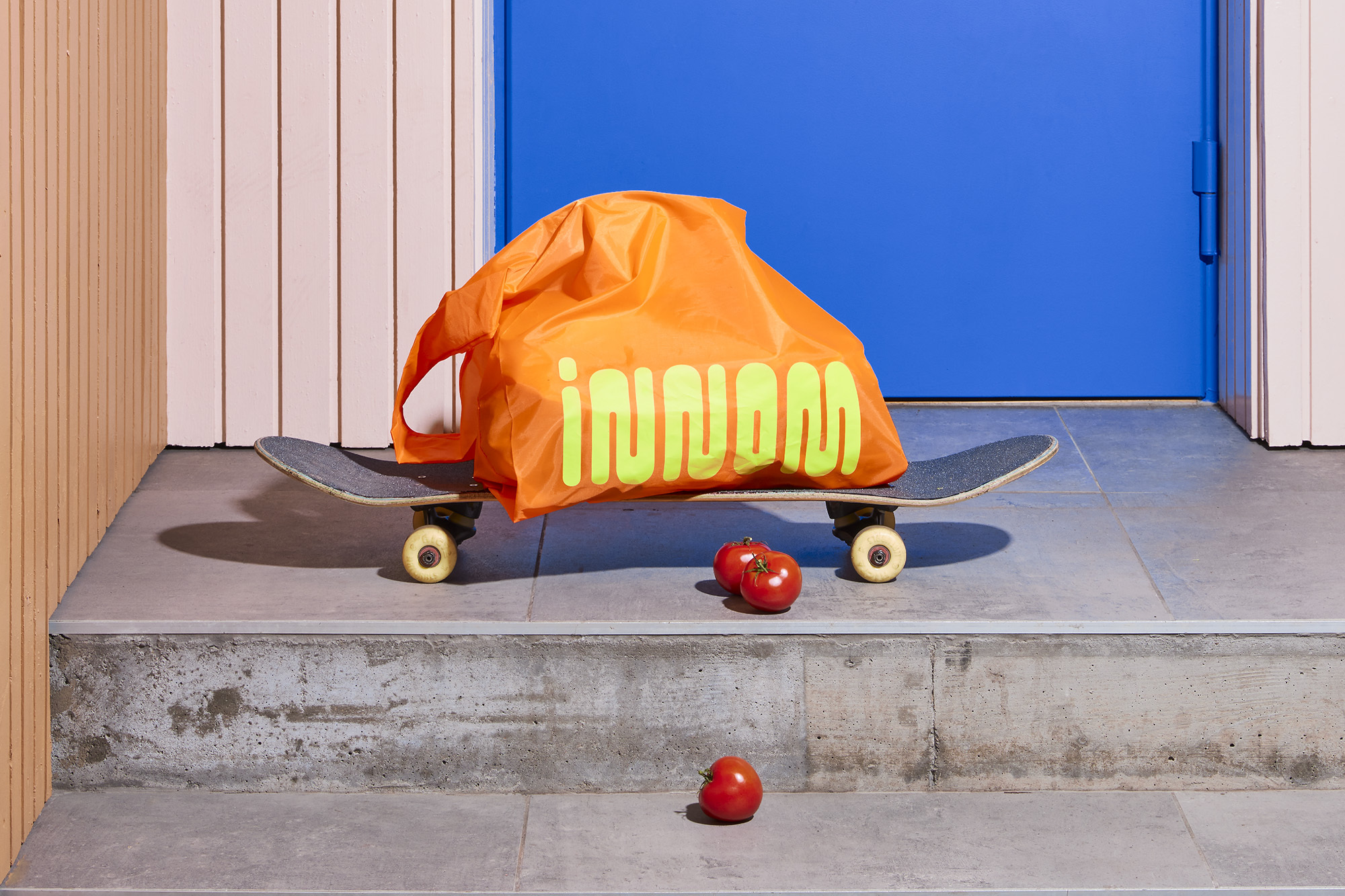 Orange reusable shopping bag on a skateboard with tomatoes on concrete steps next to a blue door and peach wall. Bright, modern aesthetic.