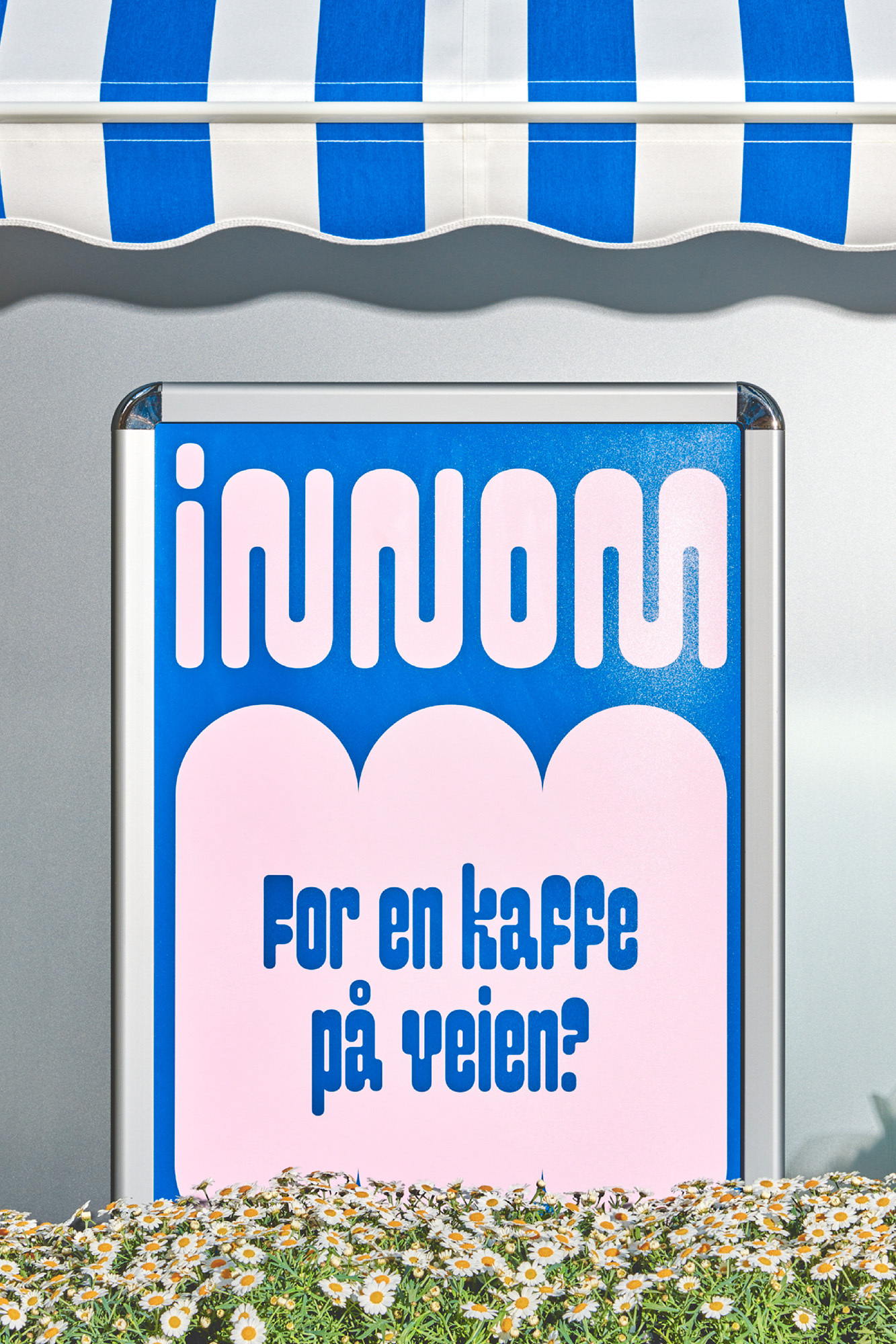 Framed sign with the text 'innom For en kaffe på veien?' sits beneath a blue and white striped awning above a flower bed of daisies.