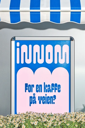 Framed sign with the text 'innom For en kaffe på veien?' sits beneath a blue and white striped awning above a flower bed of daisies.