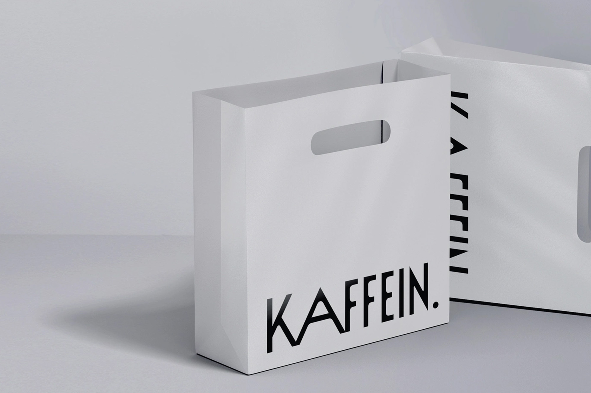 Two white paper shopping bags with black 'KAFFEIN.' branding on a soft grey background, one bag slightly behind the other.