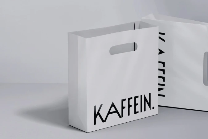 Two white paper shopping bags featuring black branding on a neutral grey and white studio background.