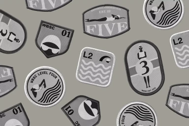An assortment of embroidered circular and shield-shaped patches with swimming and water motifs scattered on a grey background