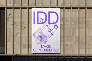 Vertical banner poster with purple graphic typography and geometric wireframe art mounted on a stone building exterior wall.