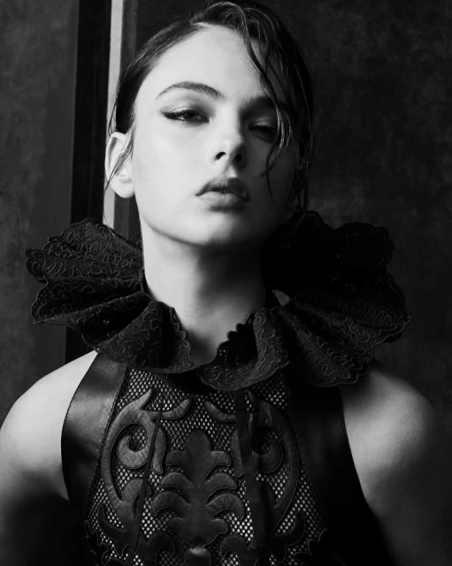 Portrait of a young woman with dark hair and striking makeup wearing an ornate black lace collar and bodice in a black-and-white photo.