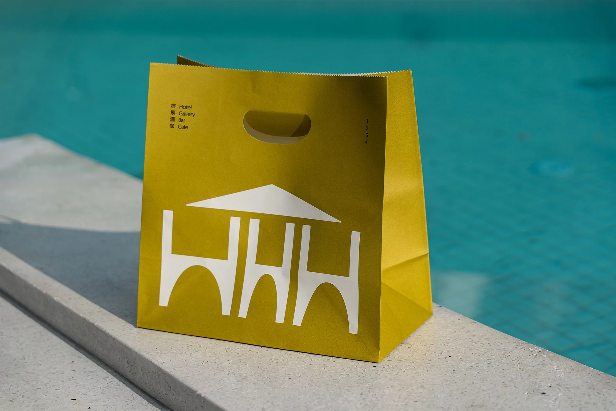 Yellow paper shopping bag with white architectural design detail, resting on a concrete ledge in front of a turquoise pool.