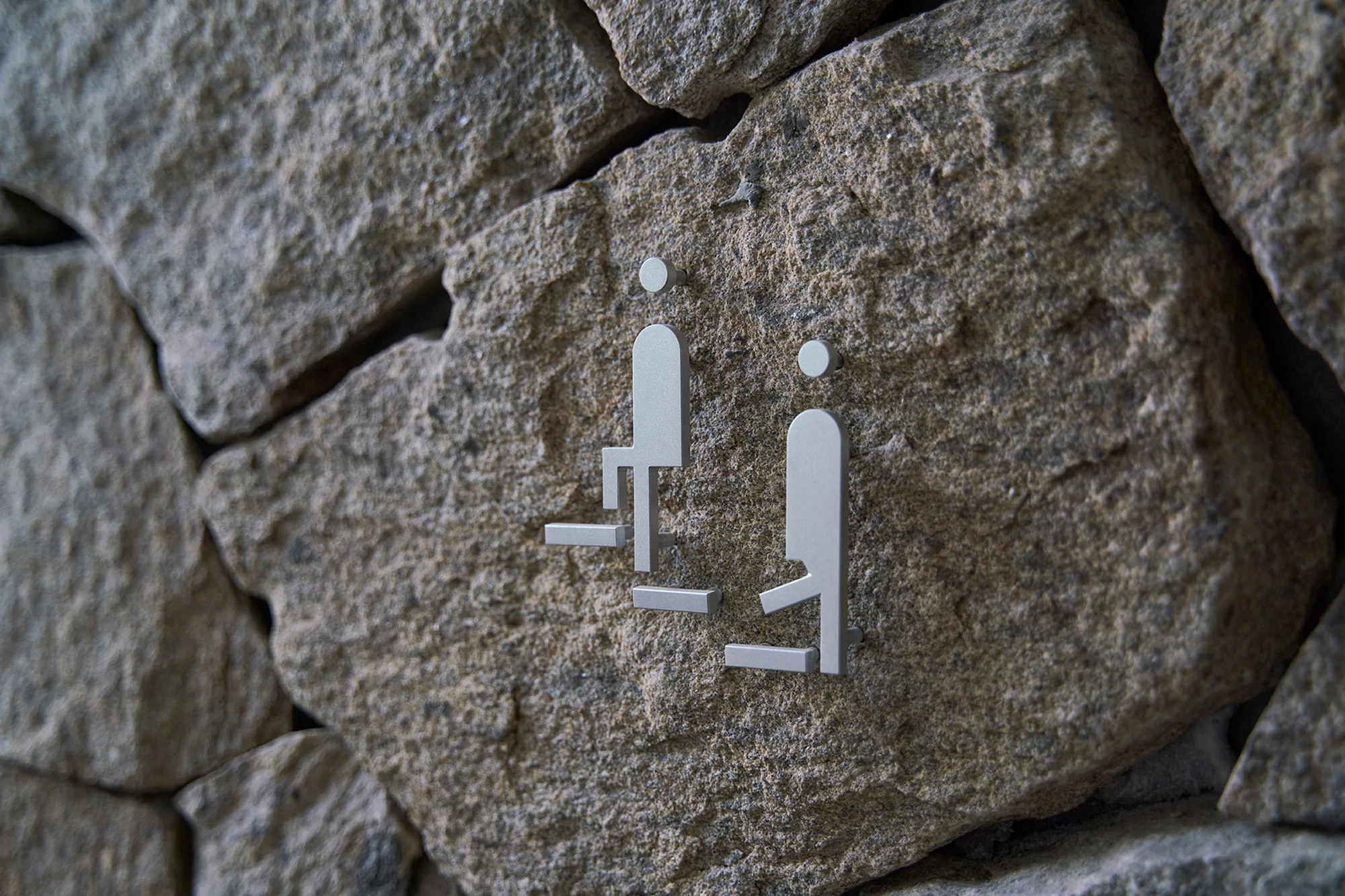 Symbolic sign on a stone wall with figures, indicating facilities for both men and women, in a close-up shot with neutral light.