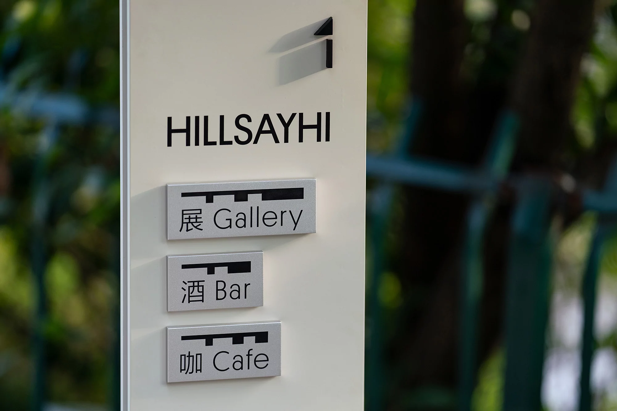 Close-up of a white sign that reads HILLSAYHI with a logo and three sets of text in Japanese and English for Gallery, Bar and Cafe.