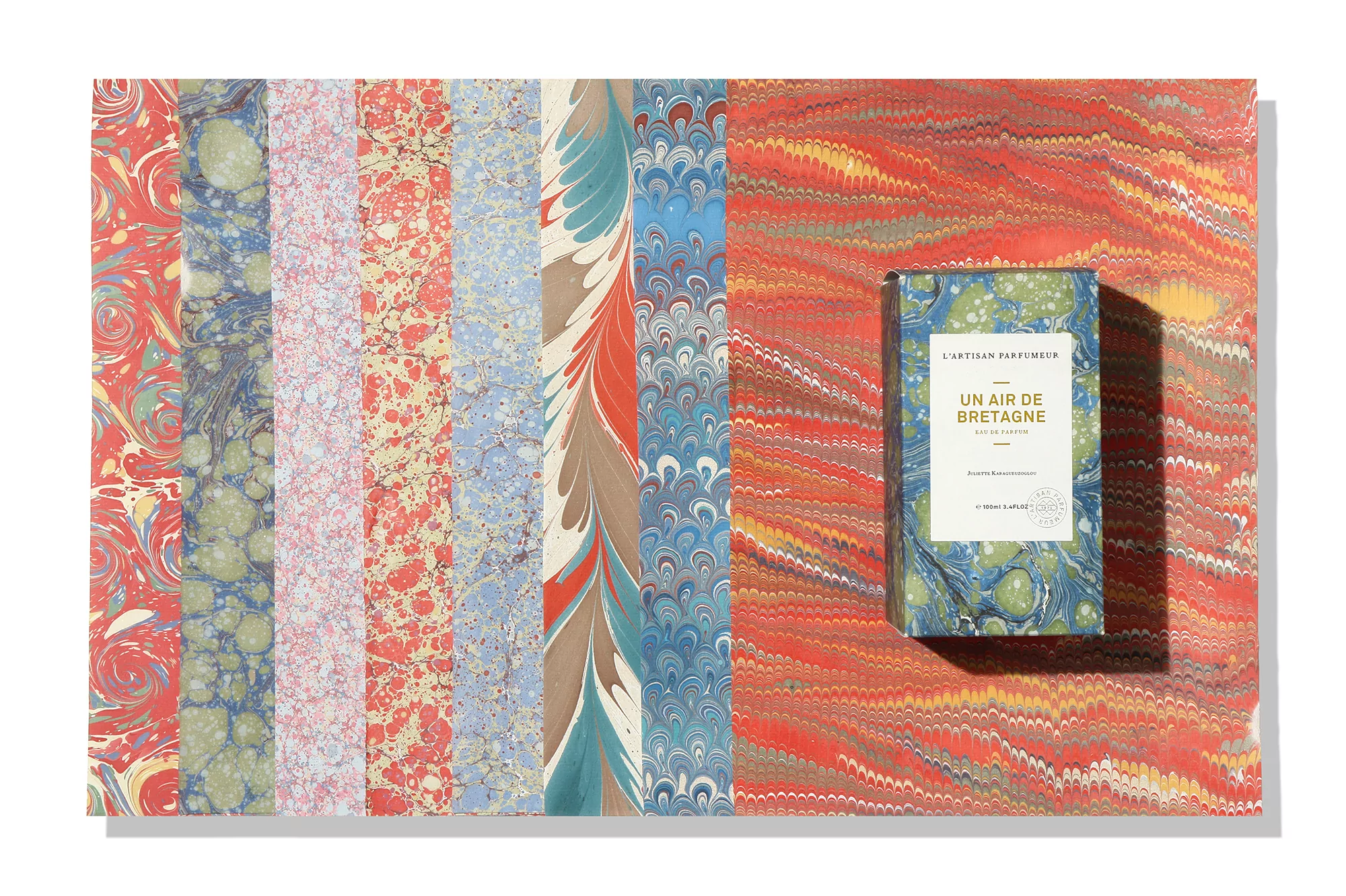 L'Artisan Parfumeur perfume box featuring a marbled design next to colorful sheets of marbled paper on a white surface.