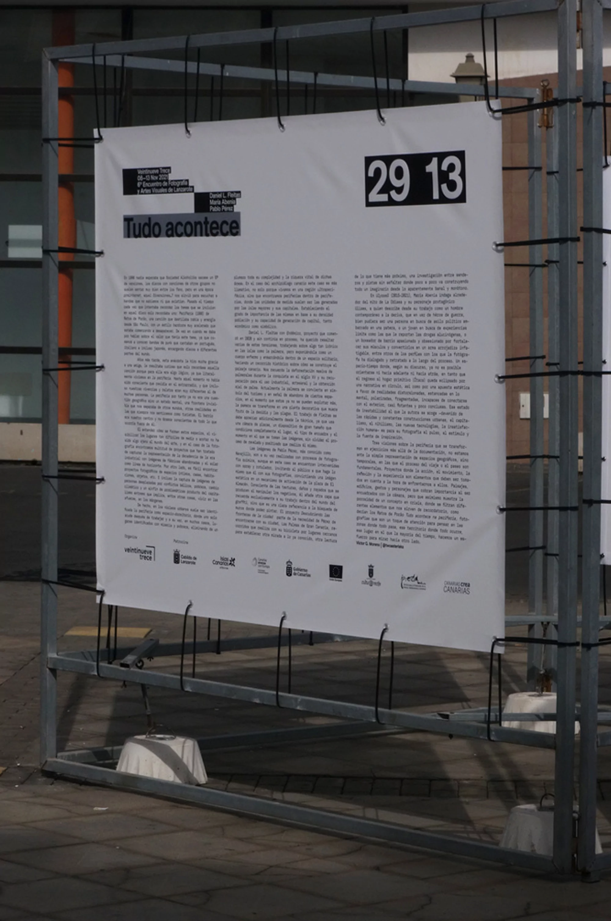 Promotional banner on a metal frame featuring black text on white background, with the inscription 'Tudo acontece' and a date '29 13'.