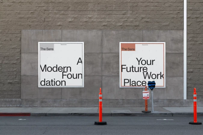 Two white posters with black text on a gray wall in an urban setting, promoting 'The Sans' with modern design and placement concepts.