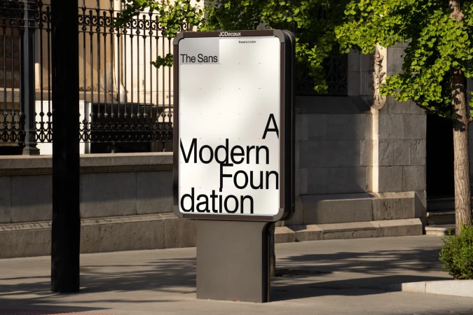 Outdoor advertising billboard on a city sidewalk, featuring the text 'The Sans, A Modern Foundation' with trees in the background.