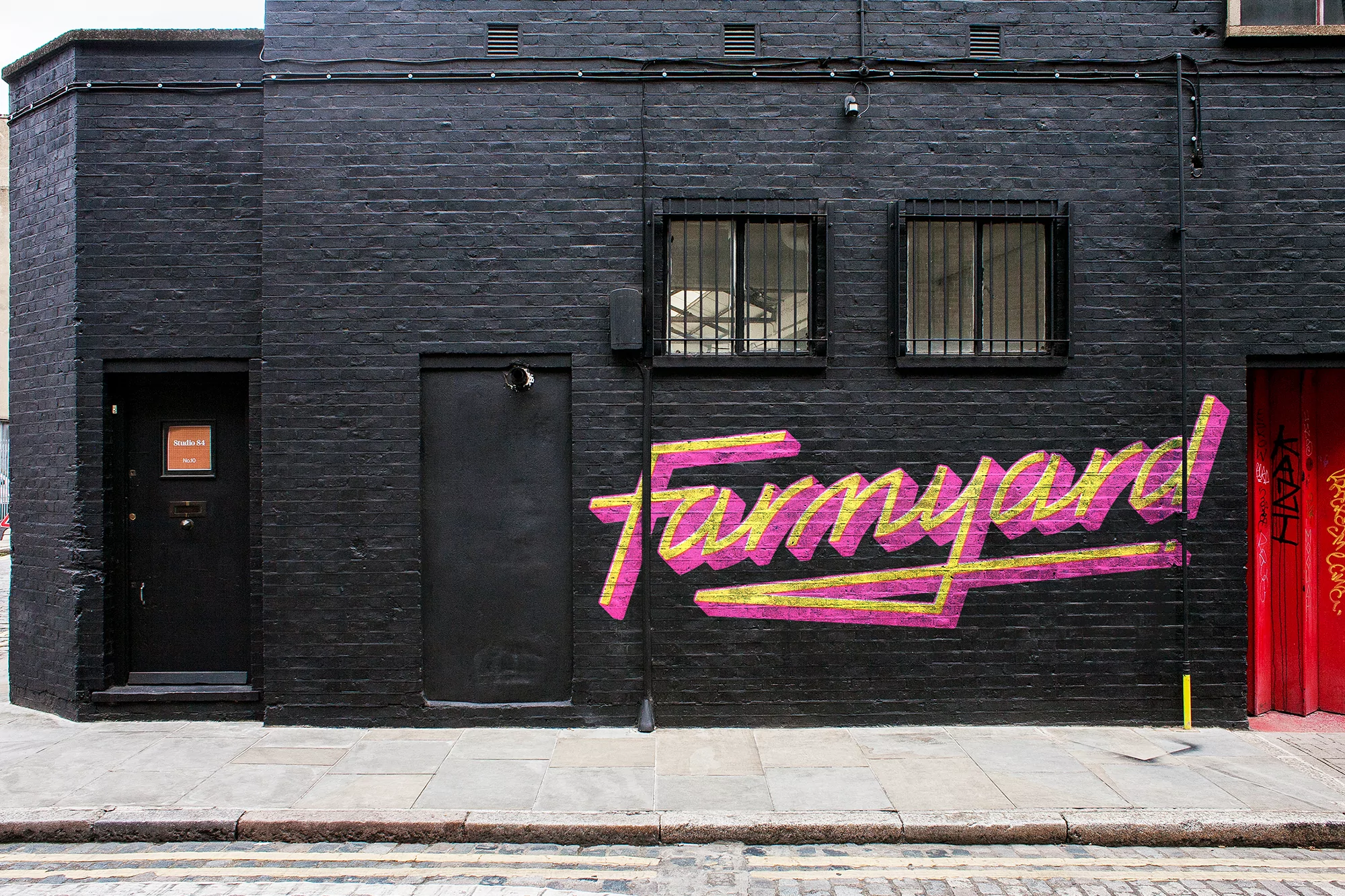 Black brick building with painted 'Farmyard' graffiti in pink and yellow, windows with bars, a black door, and a red door.