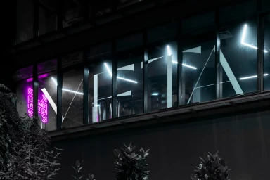 Exterior night view of a modern building with large glass windows featuring bold white typography and a bright pink neon sign.