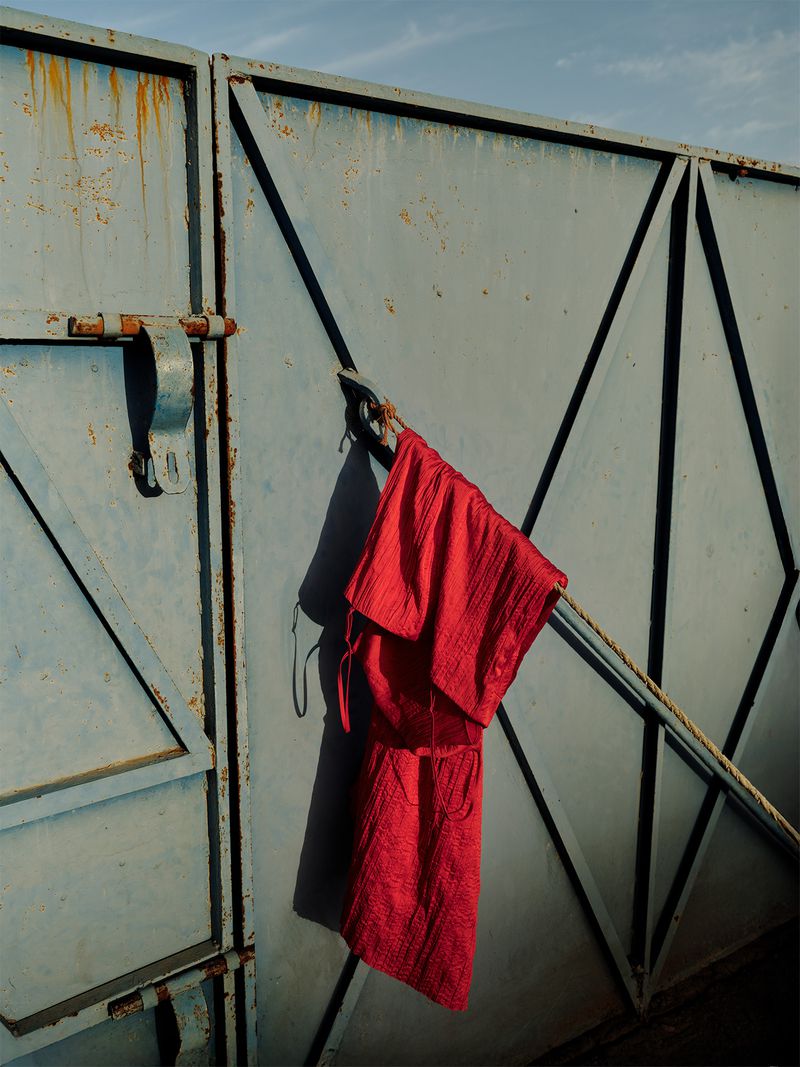 Red dress hanging on weathered blue metal gate with geometric design against a bright, partly cloudy sky, creating stark shadows.