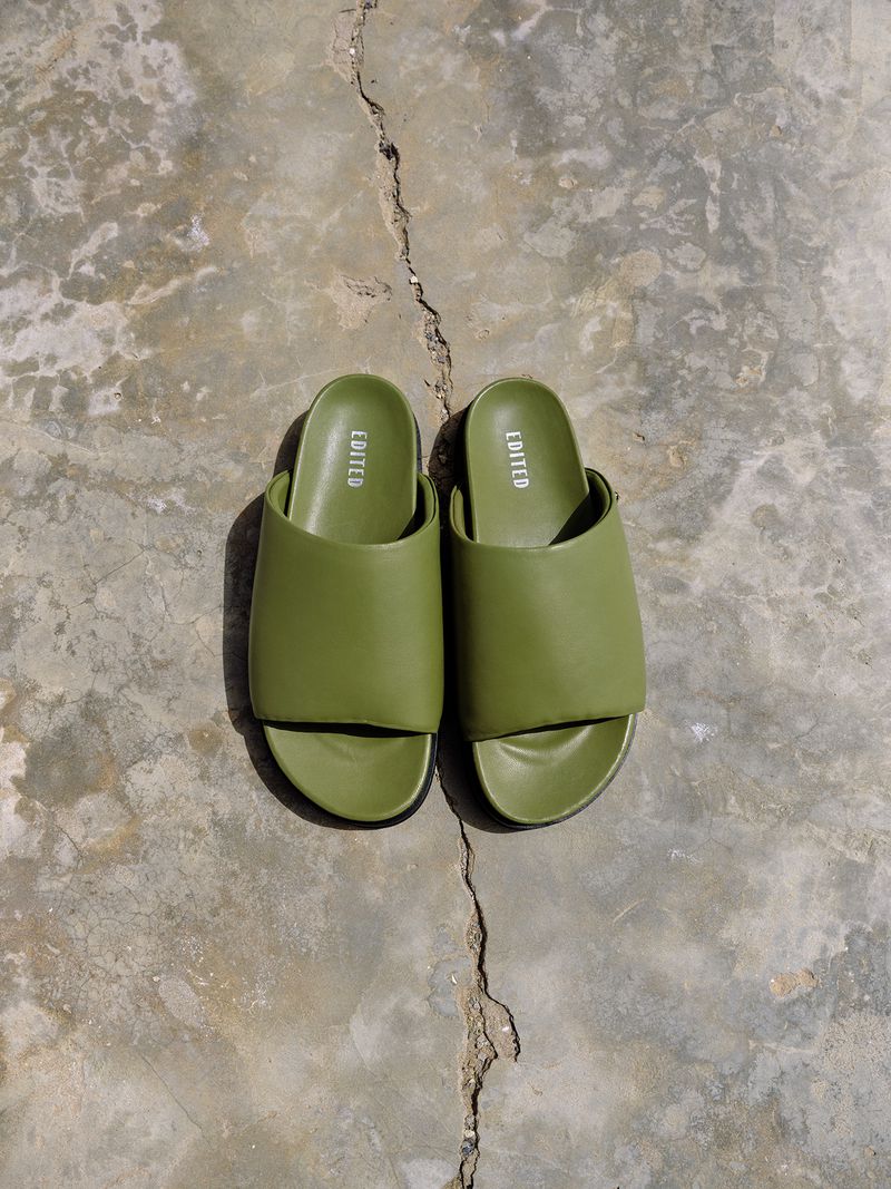 Overhead shot of olive green slide sandals on a cracked grey concrete surface with visible shadow and natural lighting. Simple fashion.